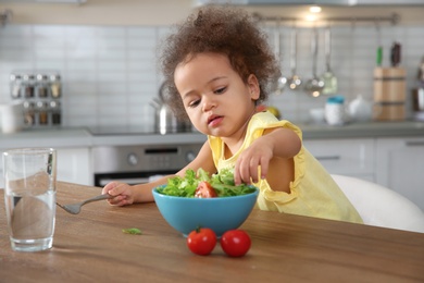 Photo of Cute African-American girl eating vegetable salad at table in kitchen