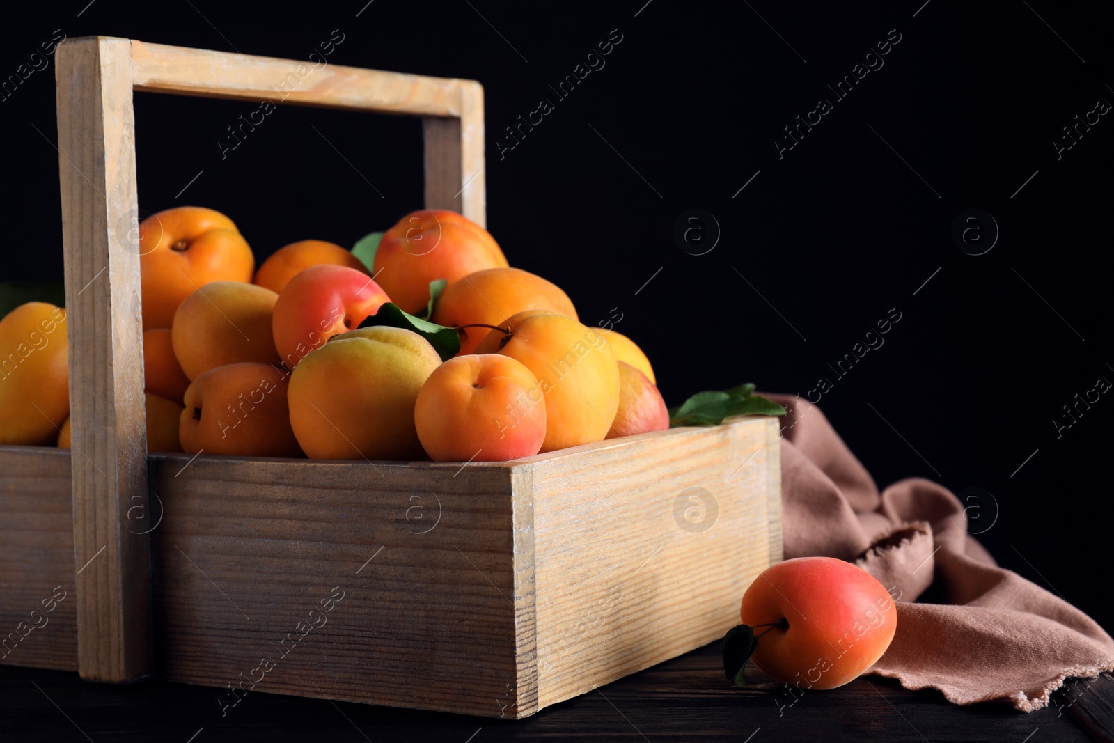 Photo of Many fresh ripe apricots in wooden basket on table against black background