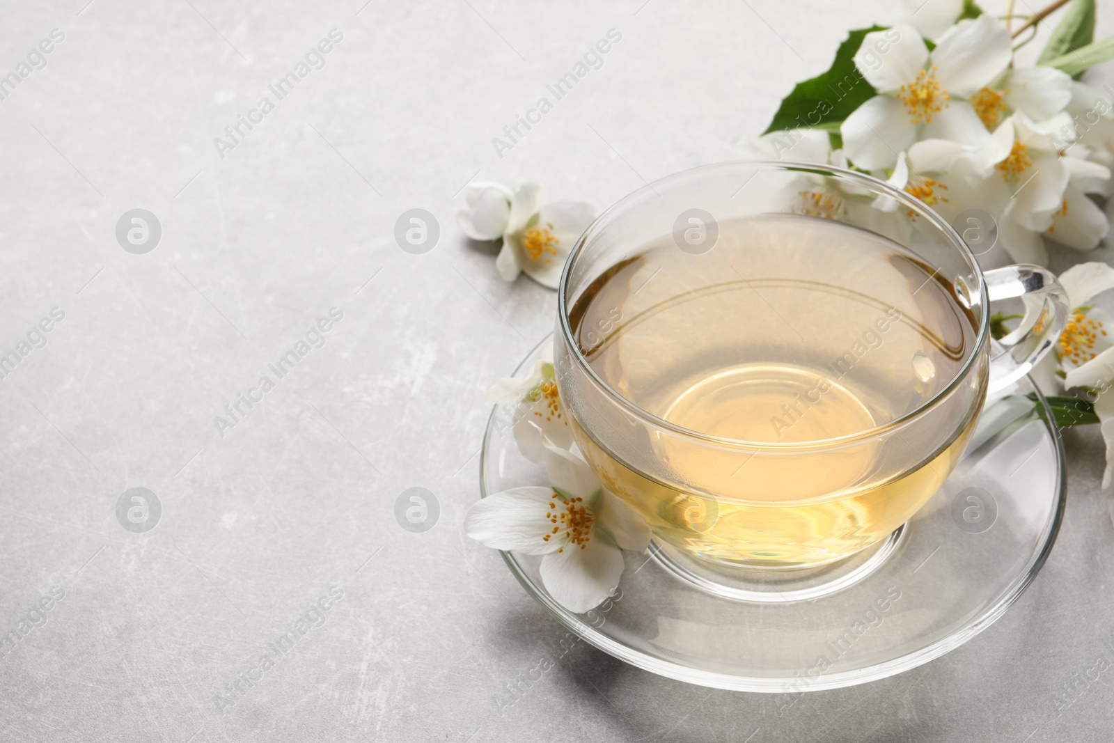 Photo of Glass cup of jasmine tea and fresh flowers on grey table. Space for text