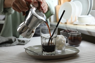 Woman pouring aromatic coffee from moka pot into glass at table in kitchen, closeup