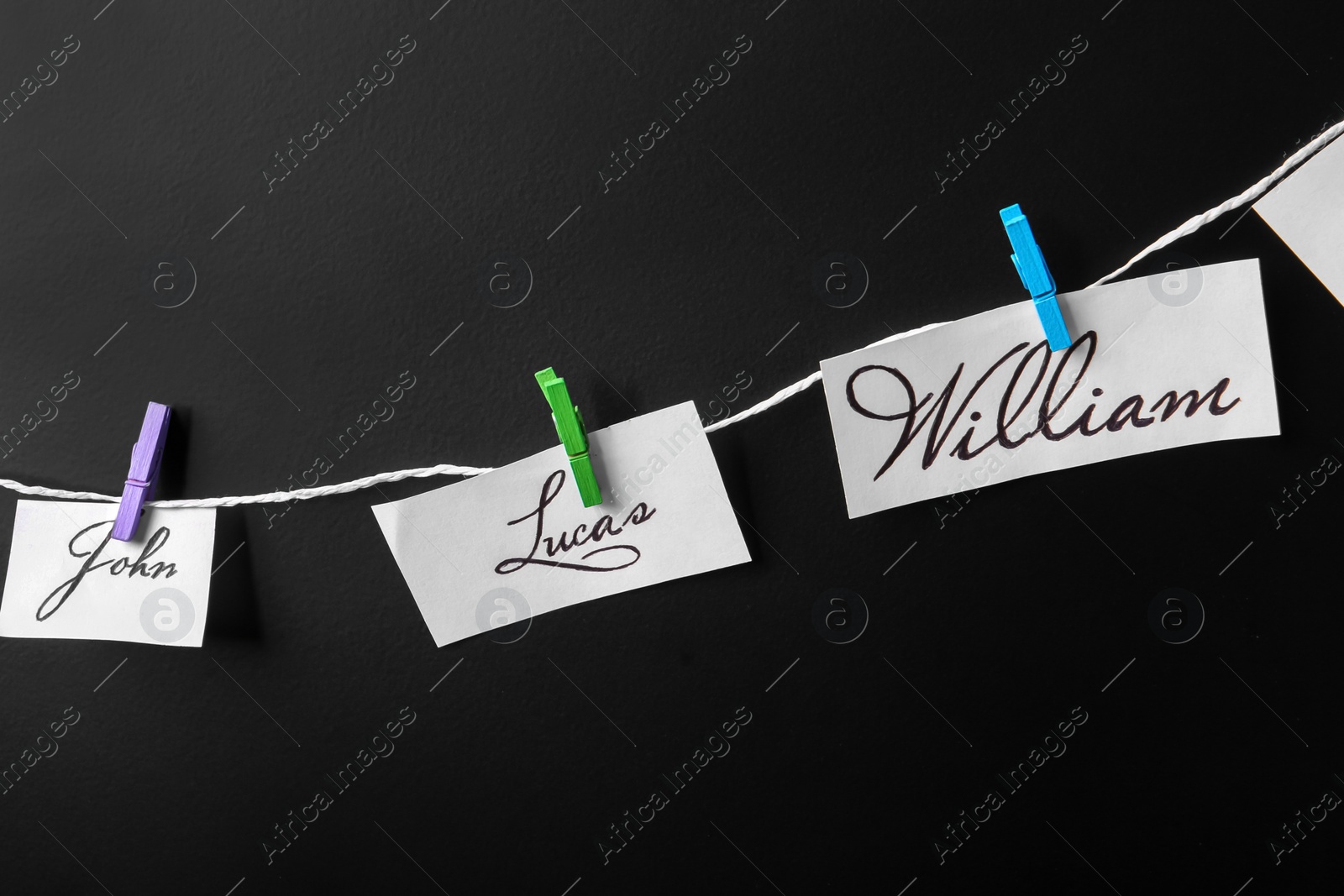 Photo of Paper pieces with baby names on rope against black background, closeup