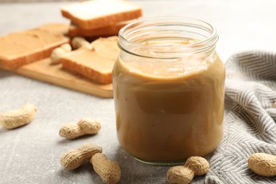 Photo of Tasty peanut butter in glass jar and peanuts on gray table, closeup