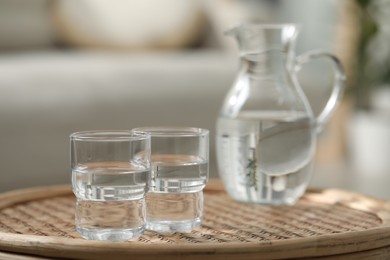 Photo of Jug and glasses with clear water on wicker surface against blurred background, closeup