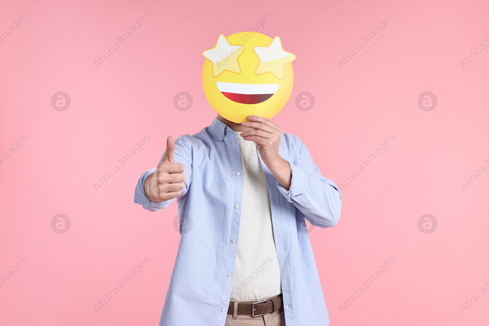 Photo of Man holding emoticon with stars instead of eyes and showing thumb up on pink background