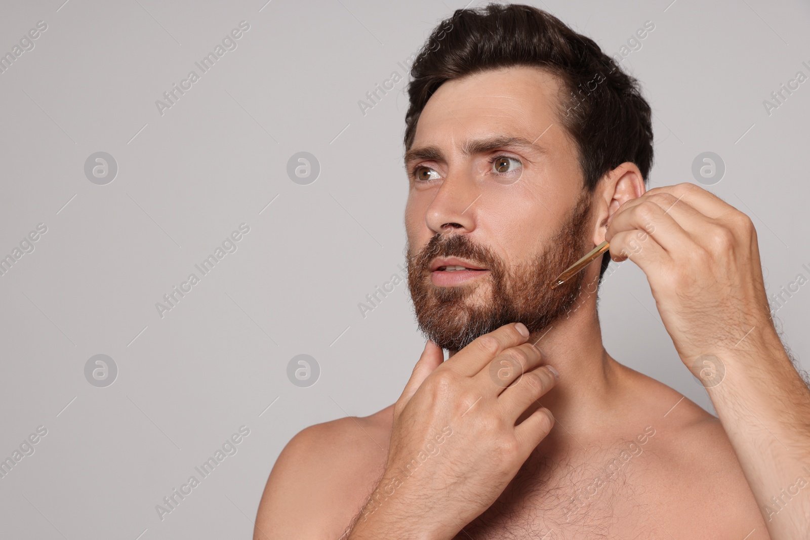 Photo of Man applying oil onto beard on grey background. Space for text