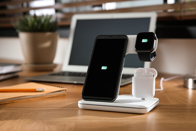 Mobile phone, earphones and smartwatch charging with wireless pad on wooden desk. Modern workplace device