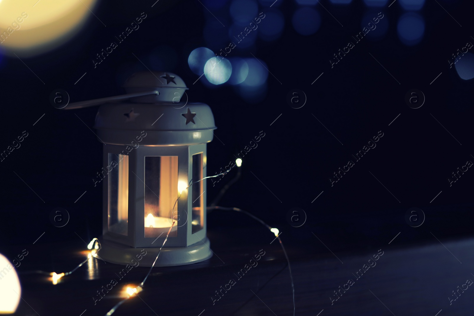 Photo of Lantern and Christmas lights on wooden railing outdoors against blurred background, space for text. Winter night