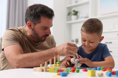 Photo of Motor skills development. Father and his little son playing with wooden lacing toy at white table indoors