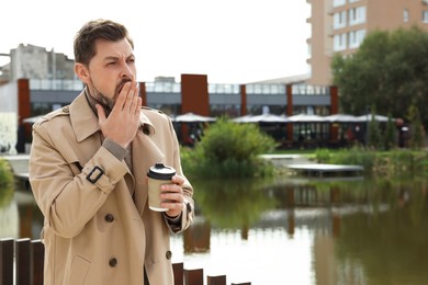 Sleepy man with cup of coffee yawning near river outdoors. Space for text