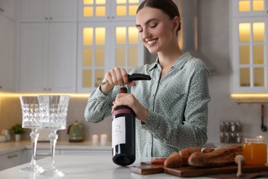 Photo of Romantic dinner. Happy woman opening wine bottle with corkscrew at table in kitchen