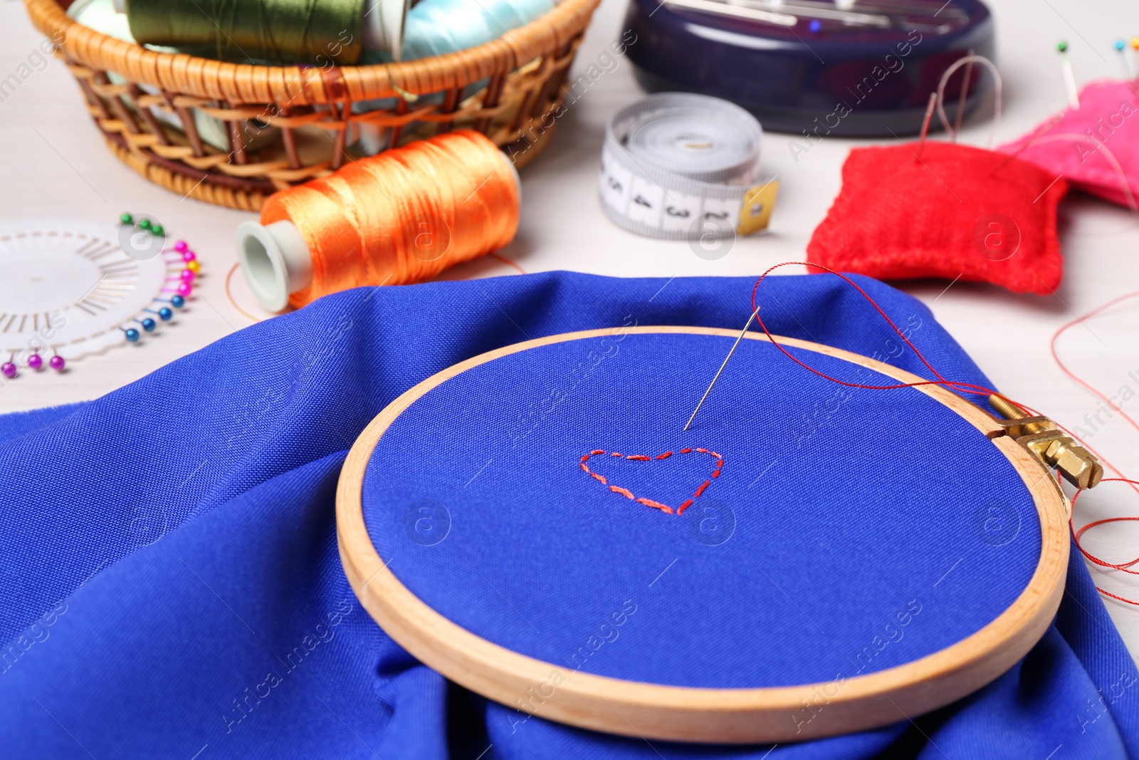 Photo of Red embroidered heart, thread and sewing needle on blue cloth with hoop on light table, closeup