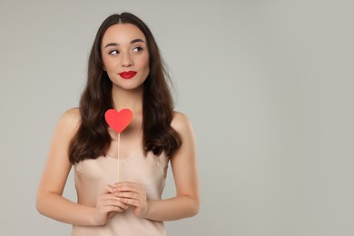 Beautiful young woman with paper heart on grey background, space for text