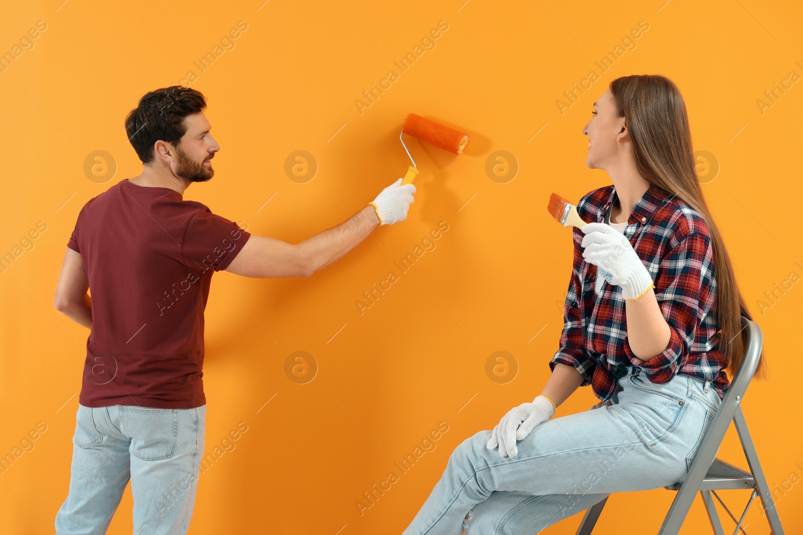 Photo of Man painting orange wall and woman with brush. Interior design