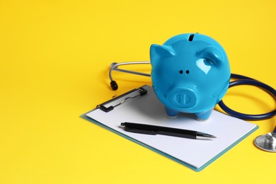 Photo of Piggy bank, stethoscope and clipboard on yellow background, space for text. Medical insurance
