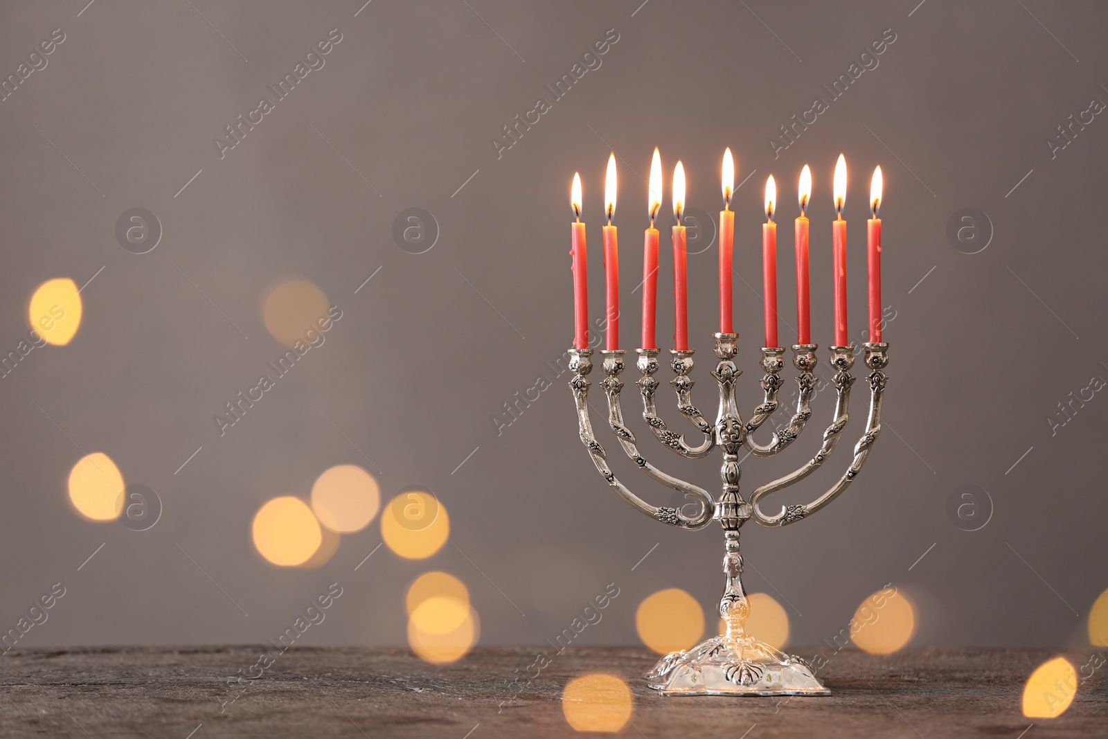 Photo of Silver menorah with burning candles on table against grey background and blurred festive lights, space for text. Hanukkah celebration