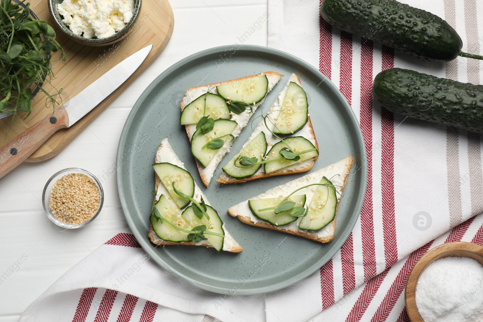 Photo of Tasty cucumber sandwiches with sesame seeds and microgreens on white wooden table, flat lay