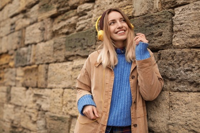 Photo of Young woman with headphones listening to music near stone wall. Space for text