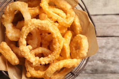 Photo of Homemade crunchy fried onion rings in wire basket on wooden background, top view