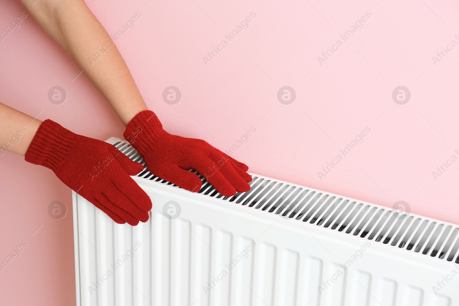 Photo of Woman in gloves warming hands on heating radiator near color wall