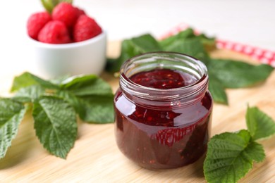 Photo of Jar of delicious raspberry jam and green leaves on wooden board, closeup. Space for text