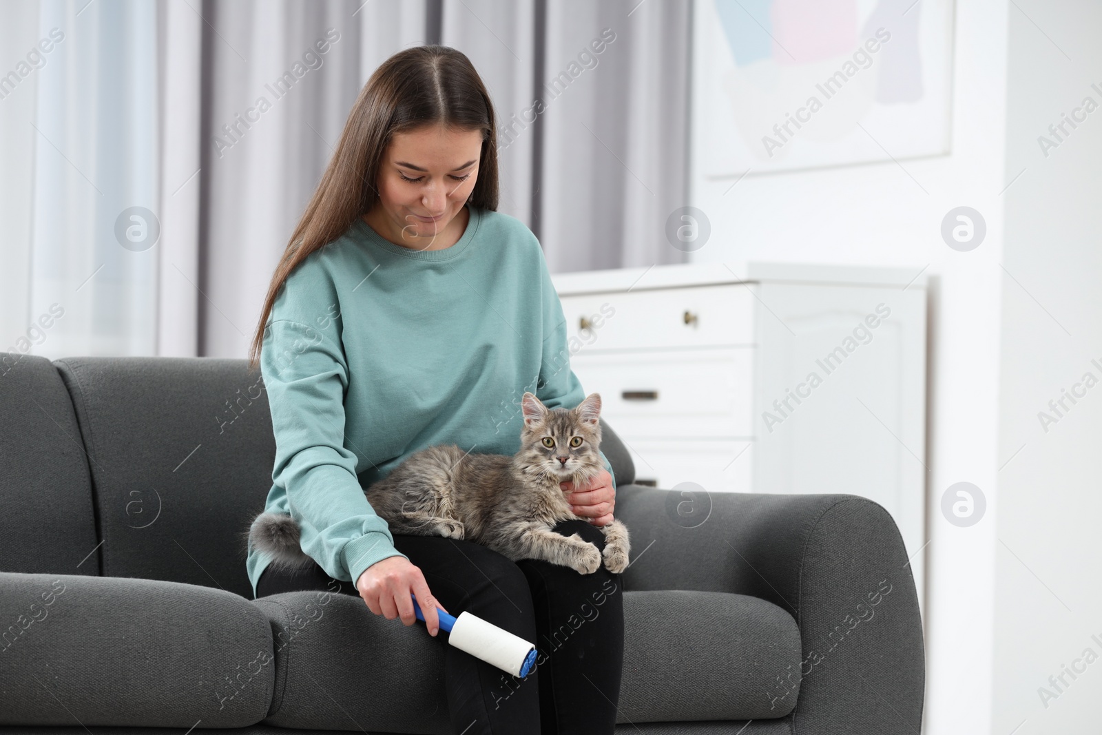 Photo of Pet shedding. Woman with lint roller removing cat`s hair from trousers on sofa at home, space for text