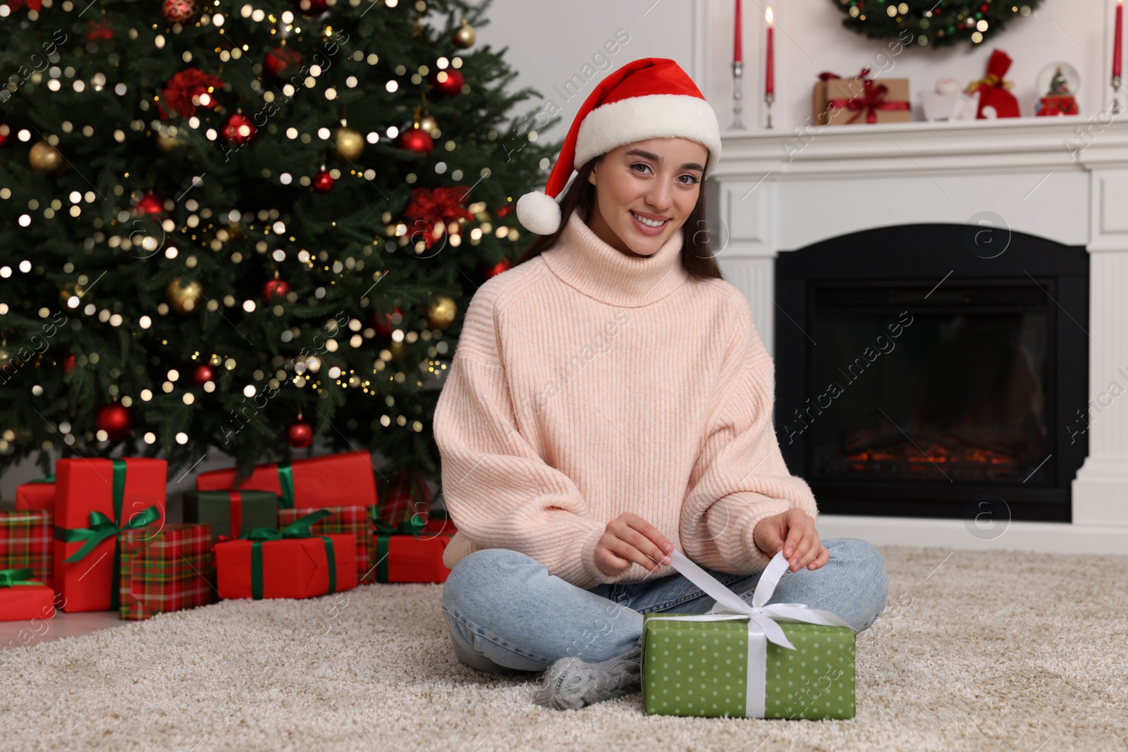 Photo of Happy young woman in Santa hat opening gift box in room decorated for Christmas