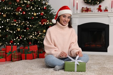 Photo of Happy young woman in Santa hat opening gift box in room decorated for Christmas