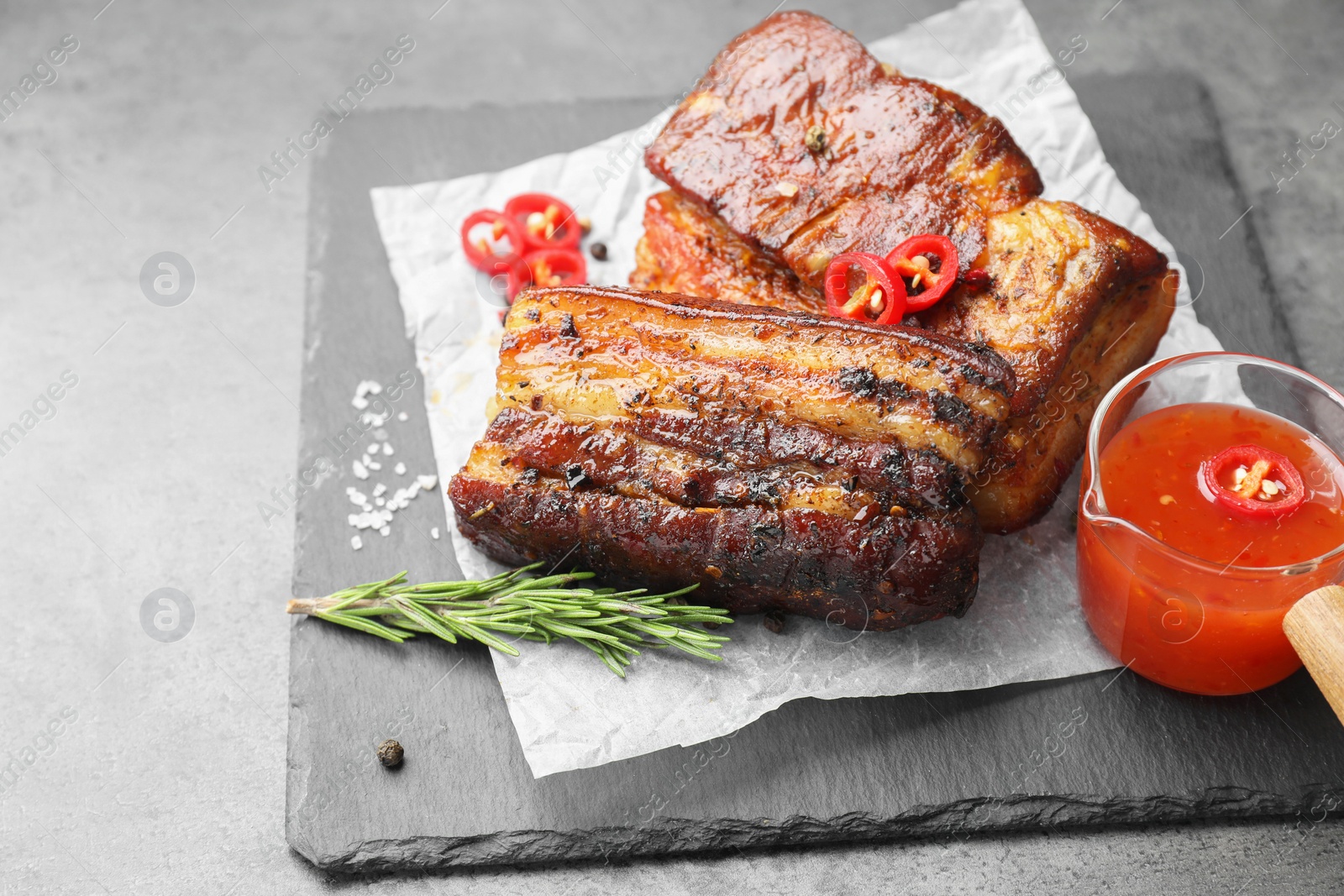 Photo of Pieces of baked pork belly served with sauce, rosemary and chili pepper on grey table