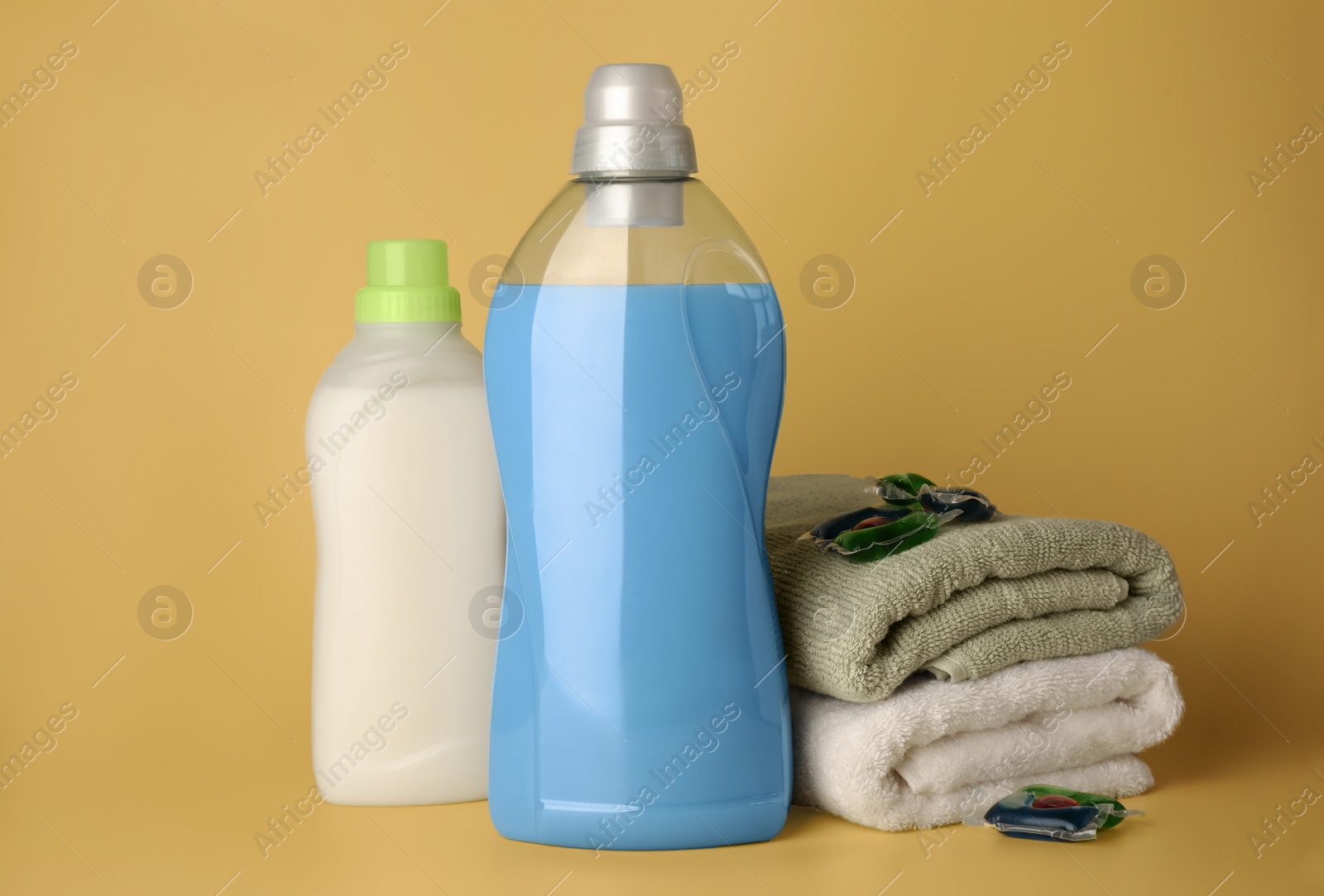 Photo of Bottles of fabric softener, laundry detergent capsules and stacked clean towels on pale yellow background