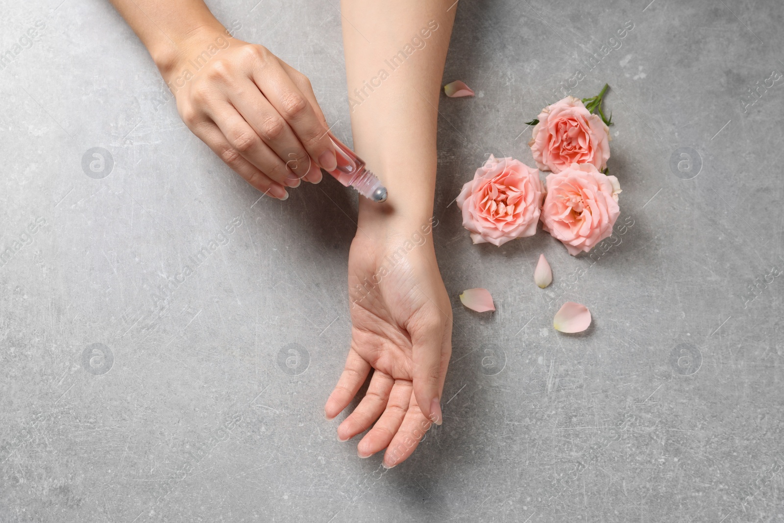 Photo of Woman applying rose essential oil on wrist and flowers at grey table, top view