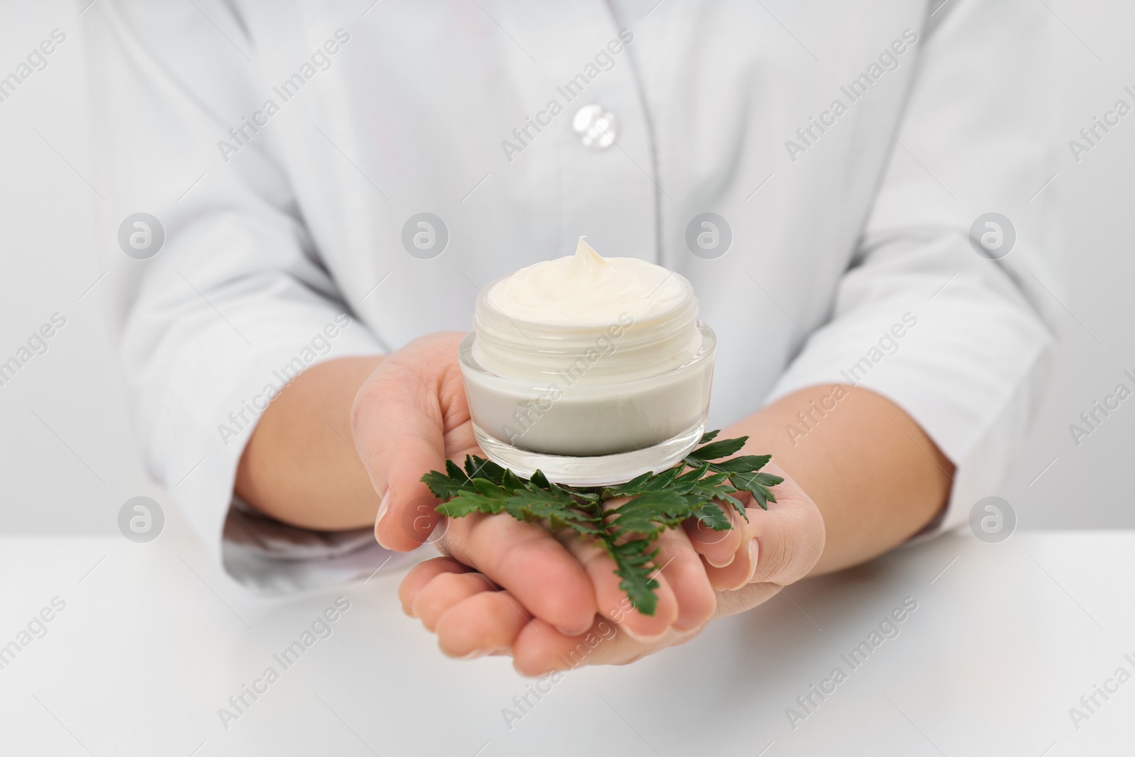 Photo of Female dermatologist holding jar of skin care product at table, closeup