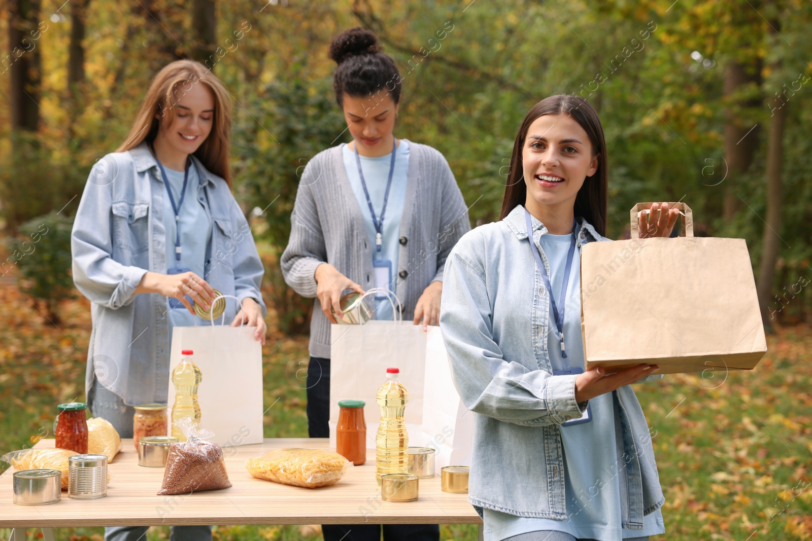 Photo of Group of volunteers packing food products at table in park. Woman with paper bag outdoors