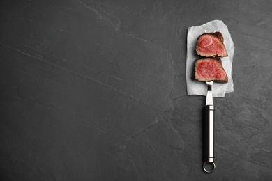 Photo of Carving fork with slices of delicious beef tenderloin on black table, top view. Space for text