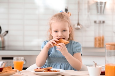 Photo of Adorable little girl eating tasty toasted bread with jam at table in kitchen