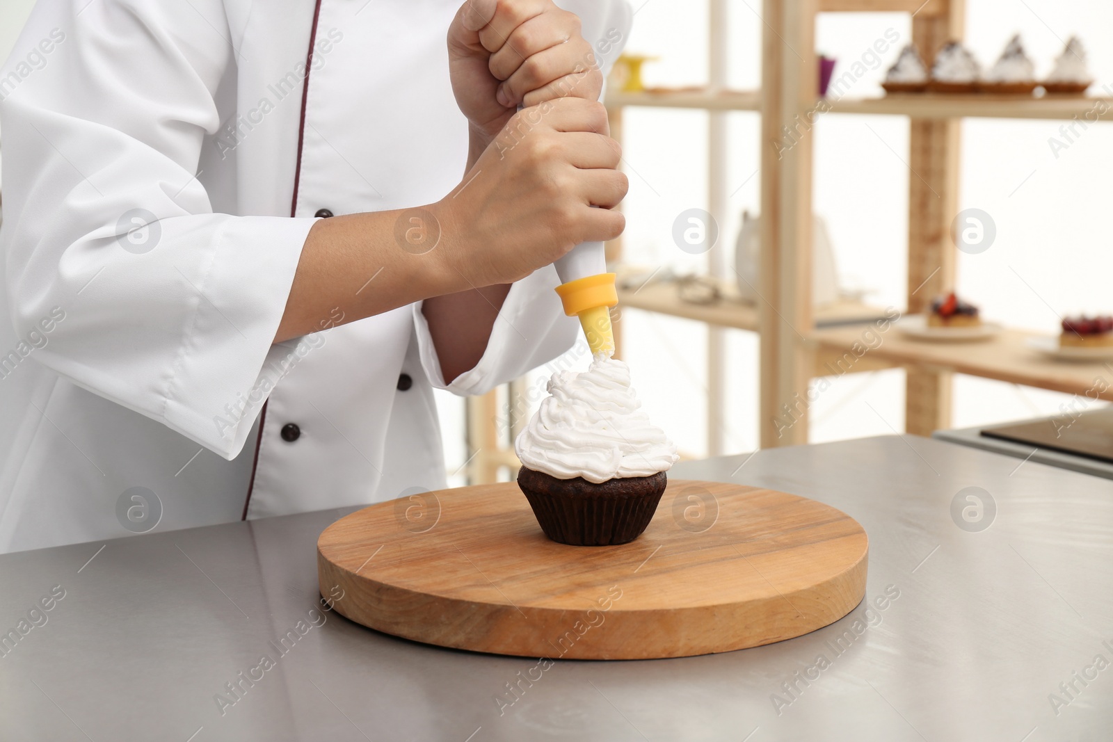 Photo of Young female pastry chef decorating cupcake with cream at table in kitchen, closeup