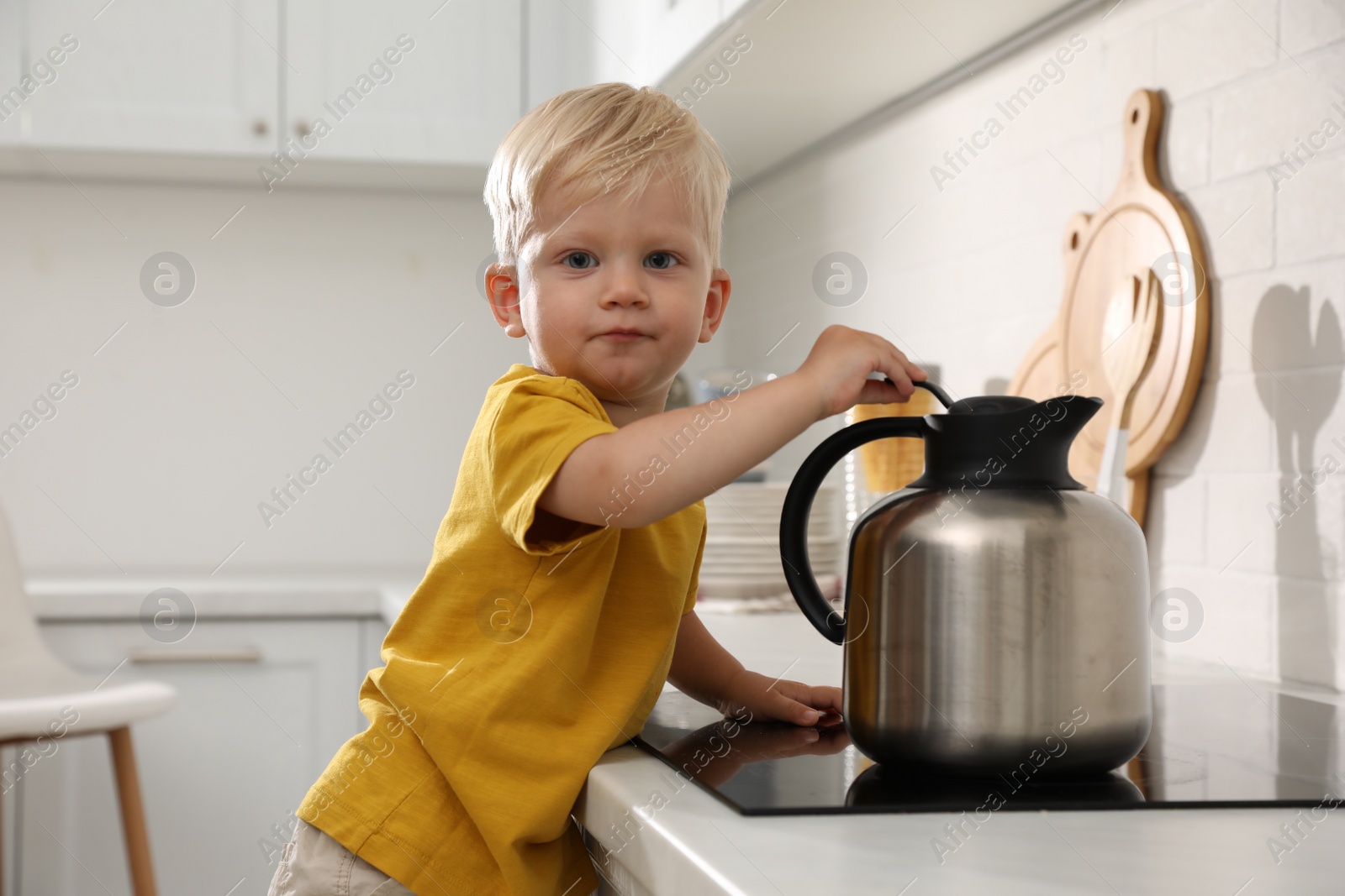 Photo of Curious little boy playing with kettle on electric stove in kitchen