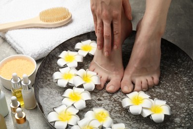 Photo of Woman soaking her feet in bowl with water and flowers on light grey floor, closeup. Spa treatment