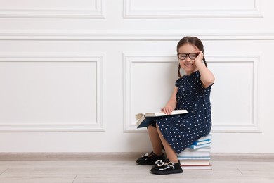 Photo of Cute little girl in glasses sitting on stack of books near white wall. Space for text