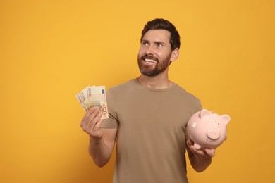 Photo of Happy man with money and ceramic piggy bank on orange background