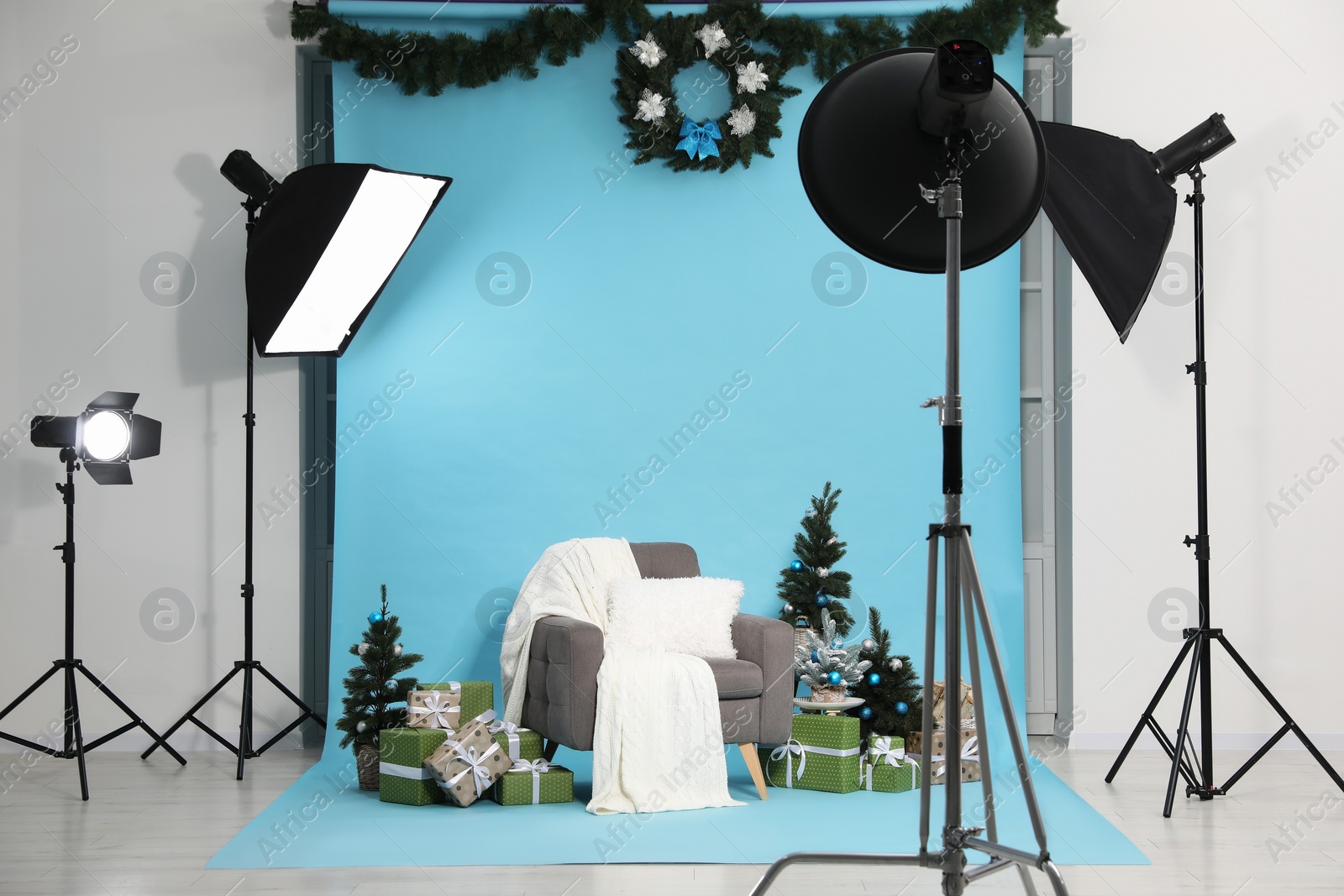 Photo of Beautiful Christmas themed photo zone with professional equipment, stylish armchair, trees and gift boxes in studio