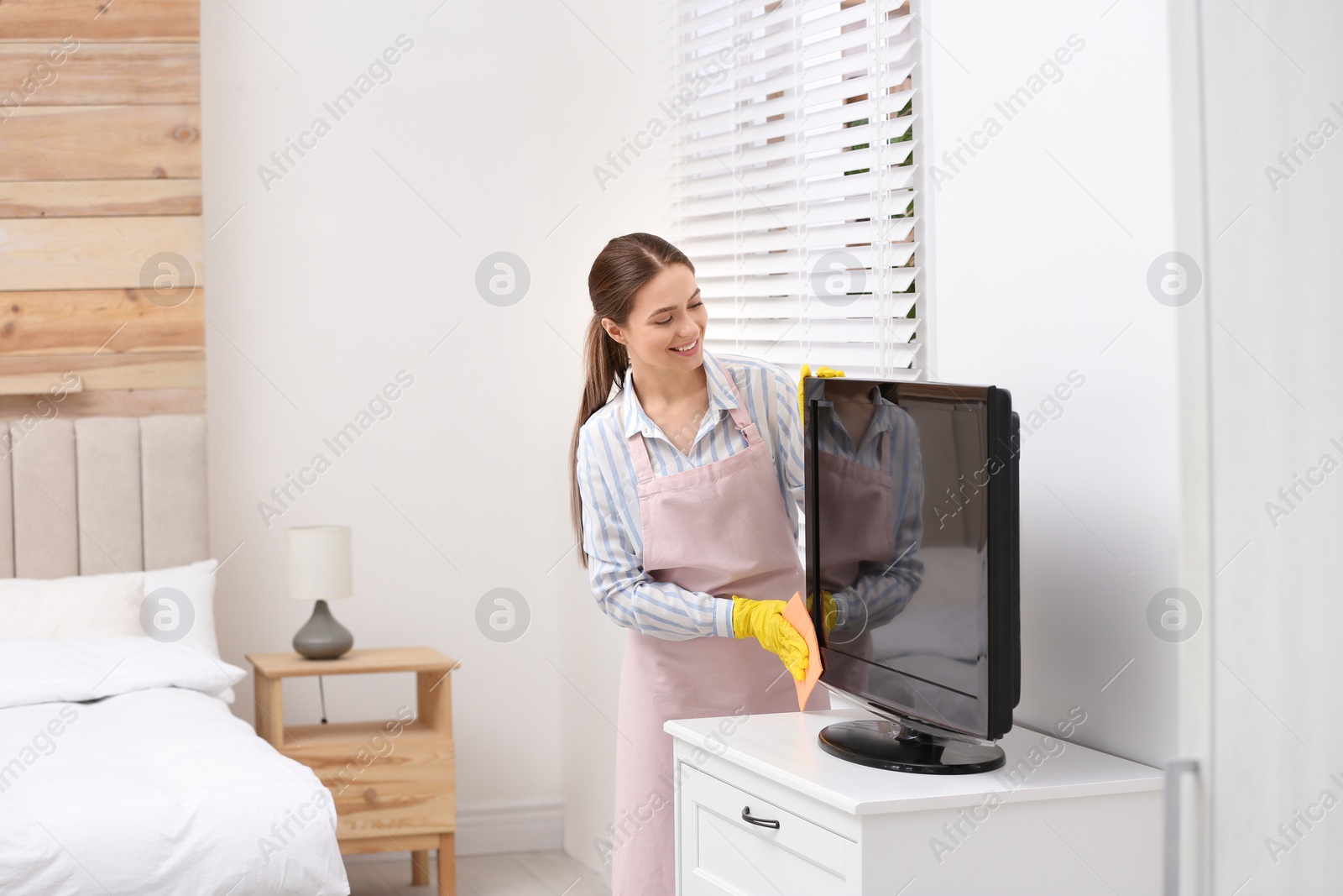 Photo of Young chambermaid wiping dust from TV with rag in hotel room