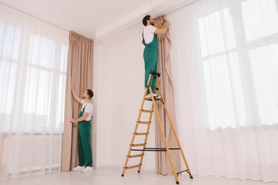 Photo of Workers in uniform hanging window curtain indoors