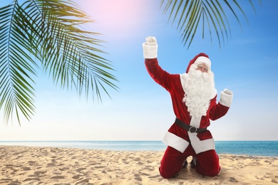 Image of Joyful Santa Claus standing on knees at sandy beach. Space for text