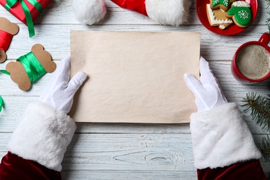 Santa Claus holding empty paper at white wooden table, top view. Space for text