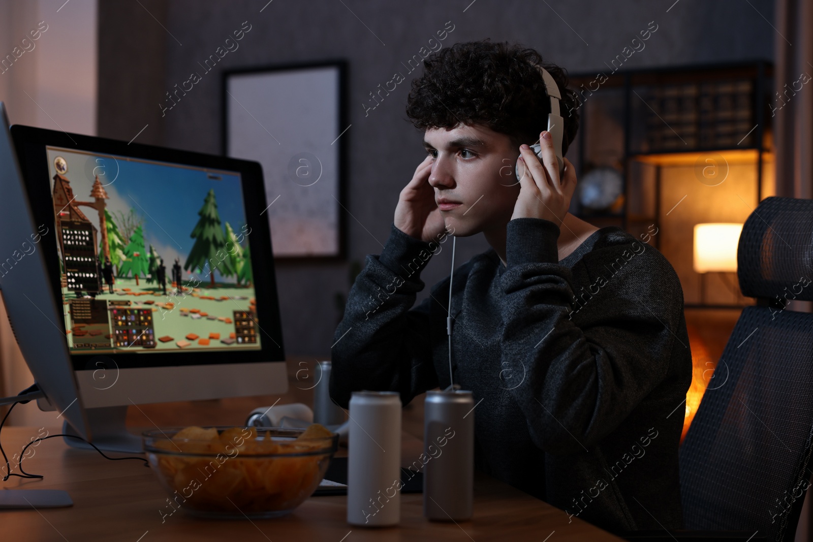 Photo of Young man with energy drink and headphones playing video game at wooden desk indoors