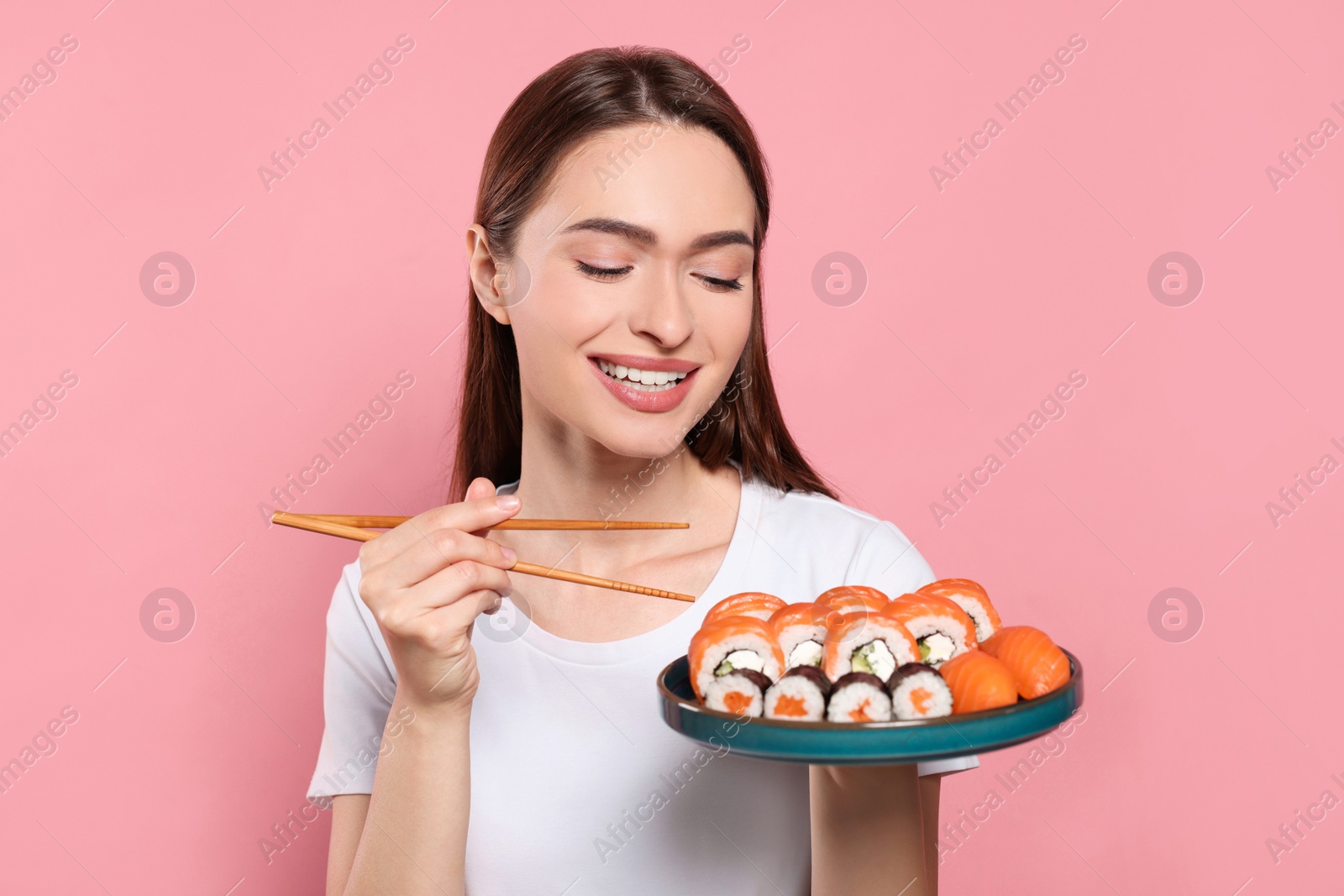 Photo of Happy young woman with plate of sushi rolls and chopsticks on pink background