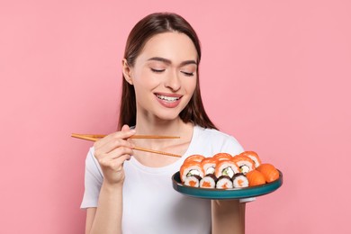 Happy young woman with plate of sushi rolls and chopsticks on pink background