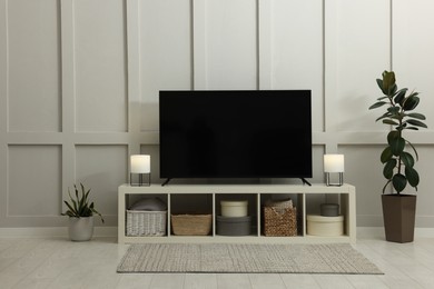 Modern TV on cabinet, lamps and beautiful houseplants near white wall in room. Interior design