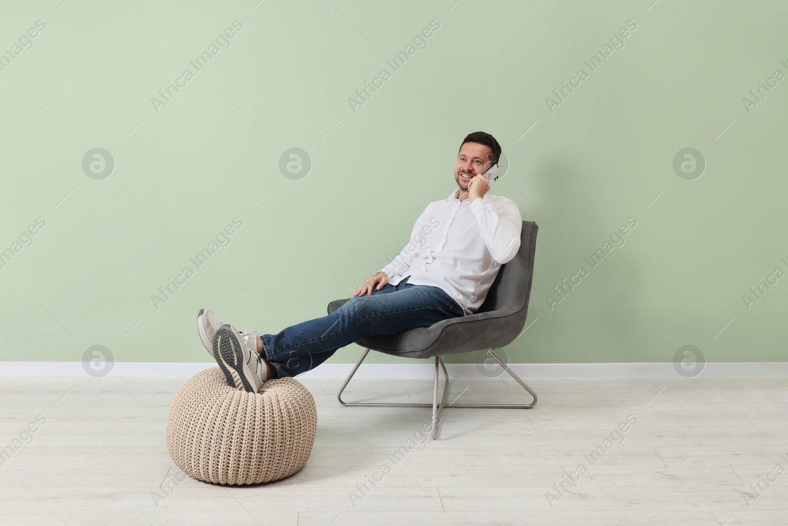 Photo of Happy man sitting in armchair and talking on smartphone near light green wall indoors