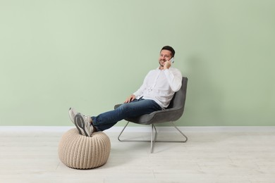 Happy man sitting in armchair and talking on smartphone near light green wall indoors